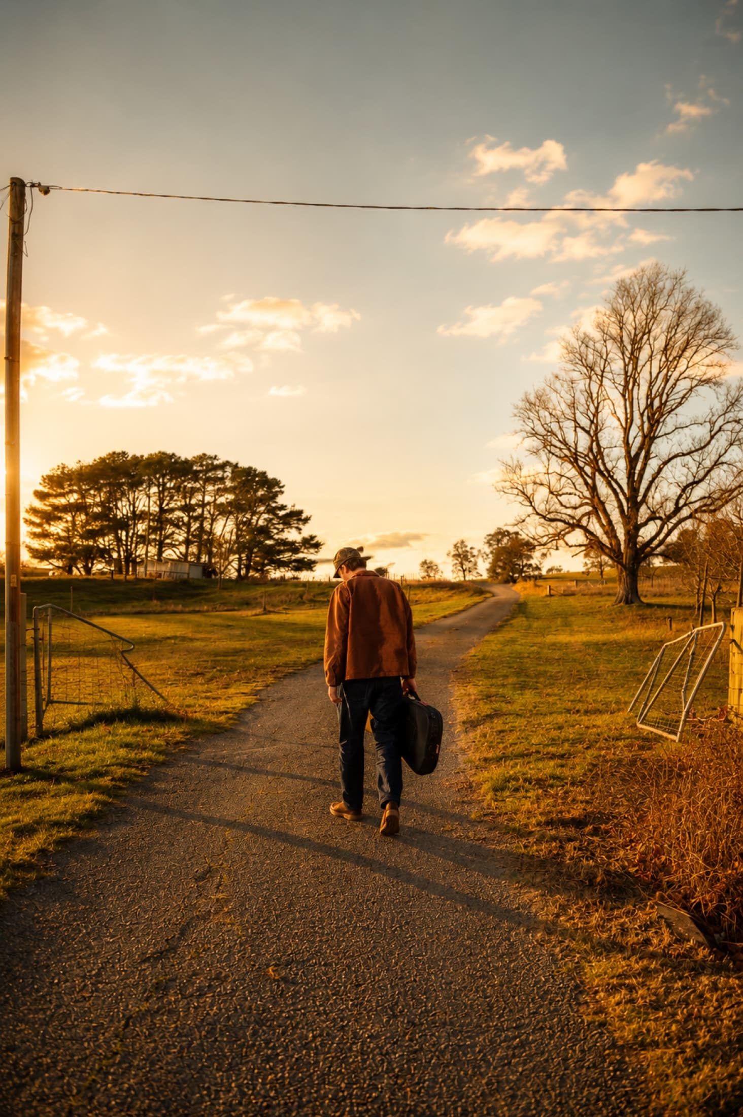 Kai Rennie walking up a country road at golden hour, guitar case in hand